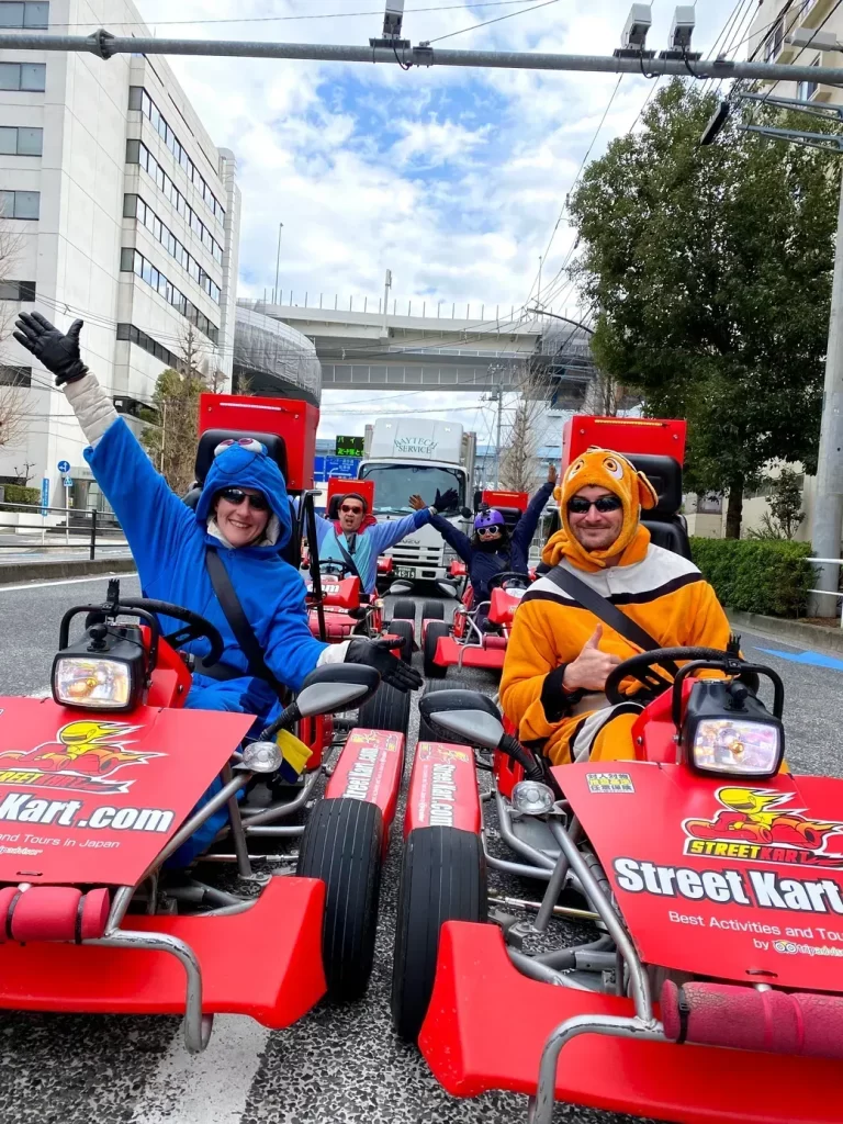 Group of people in red Street Kart go-karts wearing colorful animal costumes (blue dragon and orange tiger) waving and smiling on a city street.