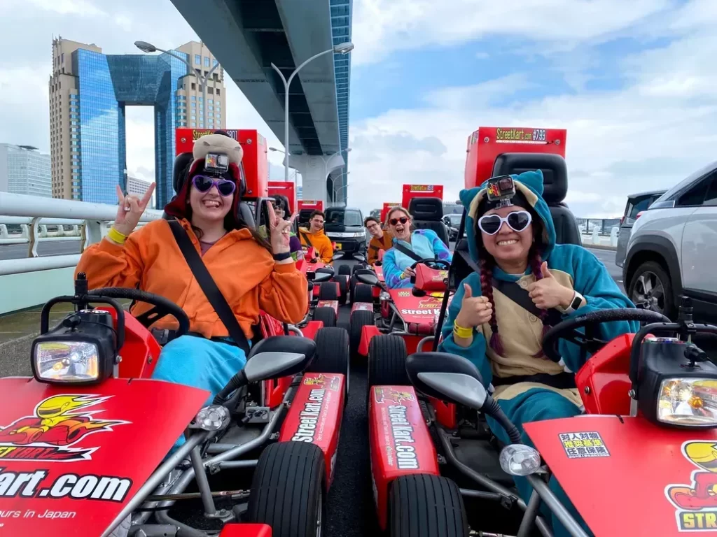 Group of friends in colorful outfits on red go-karts, posing with peace signs under an elevated roadway, city skyline in the background, GoPro cameras on helmets.