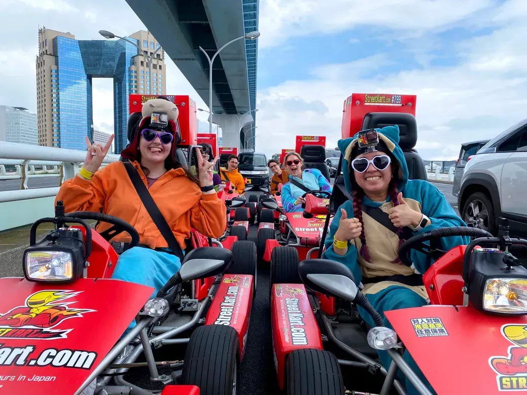 Group of friends in colorful outfits on red go-karts, posing with peace signs under an elevated roadway, city skyline in the background, GoPro cameras on helmets.