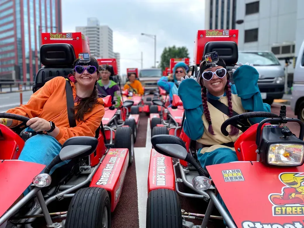 Group of friends in red go-karts lined up on a city street, smiling at the camera with GoPro cameras on their helmets.