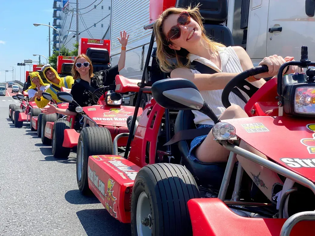 Smiling woman in sunglasses driving a red go-kart at a parade on a sunny city street, with others behind in line of carts.